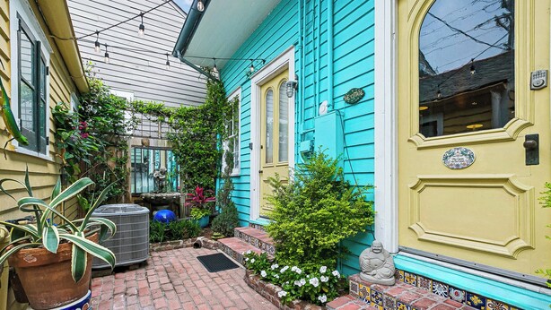 Une cour avec un chemin en briques, une maison bleue avec une porte jaune et un petit jardin agrémenté de fleurs et d’une statue.