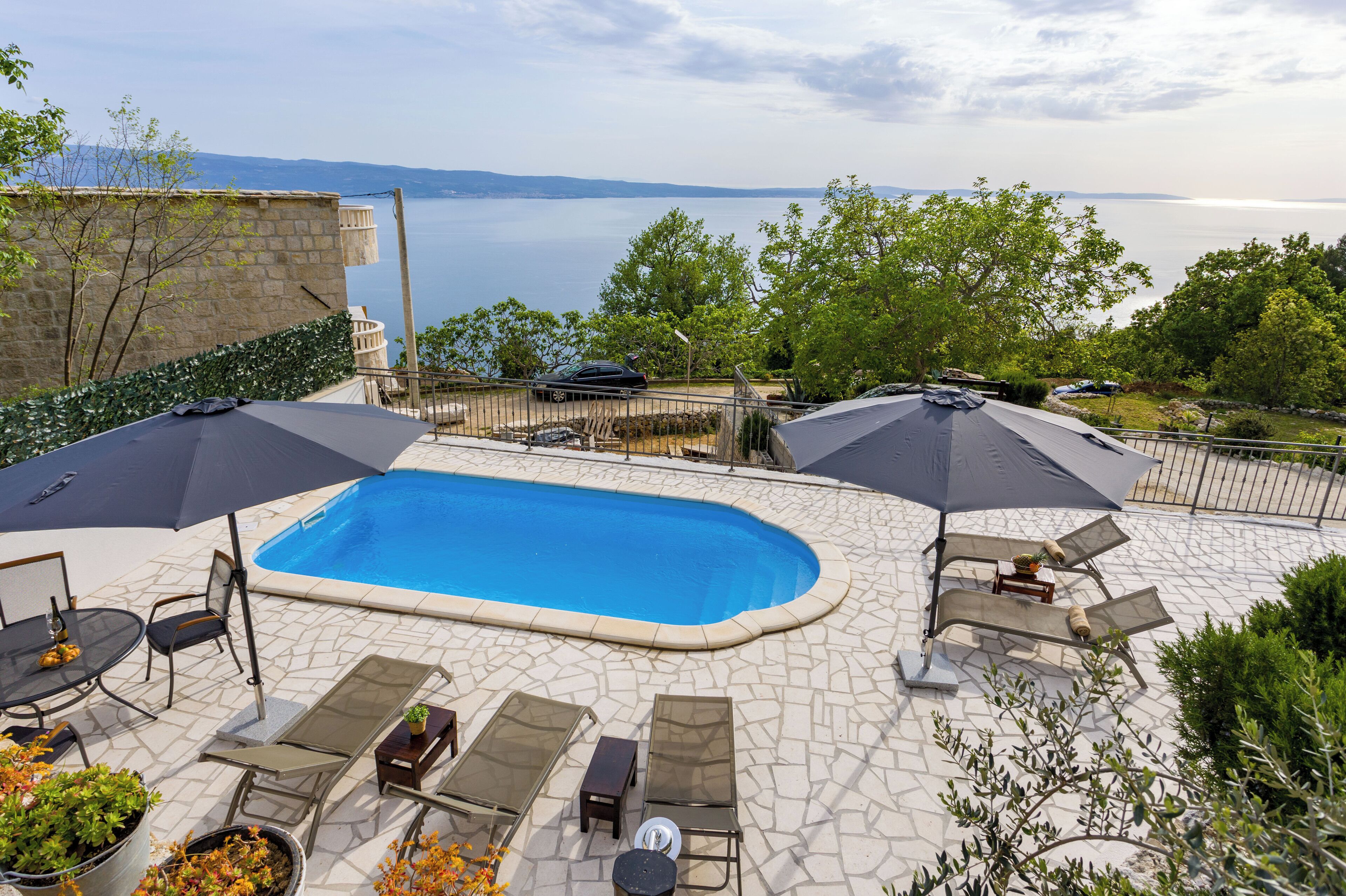 A swimming pool with lounge chairs and umbrellas, surrounded by stone tiles and greenery, overlooking a body of water.
