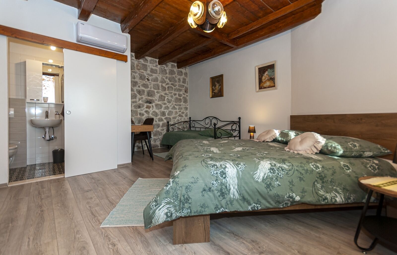 A bedroom with a wooden ceiling, a stone wall, a bed with floral bedding, a bedside table, and a desk with a chair.