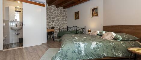 A bedroom with a wooden ceiling, a stone wall, a bed with floral bedding, a bedside table, and a desk with a chair.