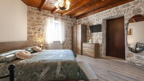 Une chambre avec un mur en pierre, des poutres en bois au plafond, un lit avec une literie à motifs floraux, une commode, un téléviseur et un miroir rond.