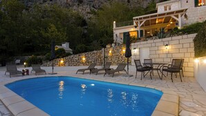 Un espace piscine agrémenté de chaises longues et de tables, entouré de murs en pierre et avec une chaîne de montagnes en toile de fond.