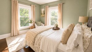 A bedroom with a large bed, two windows with curtains, a bedside table with a lamp, and a view of greenery outside. - Comfy Updated Historic Gem Pets OK 3B Vibrant Area (Painesville)