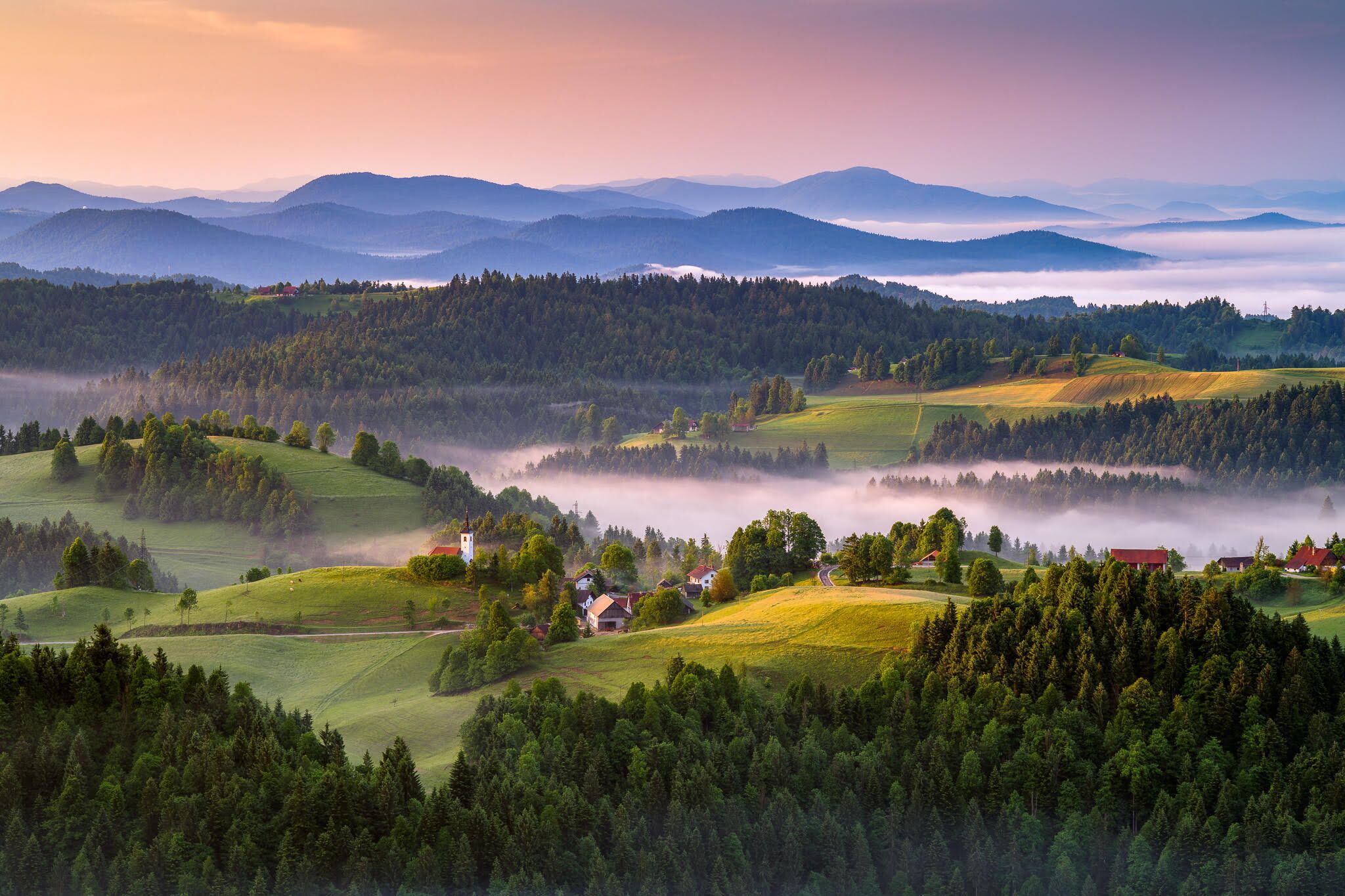 A panoramic view of a mountainous landscape with rolling green fields, a church, and scattered houses.
