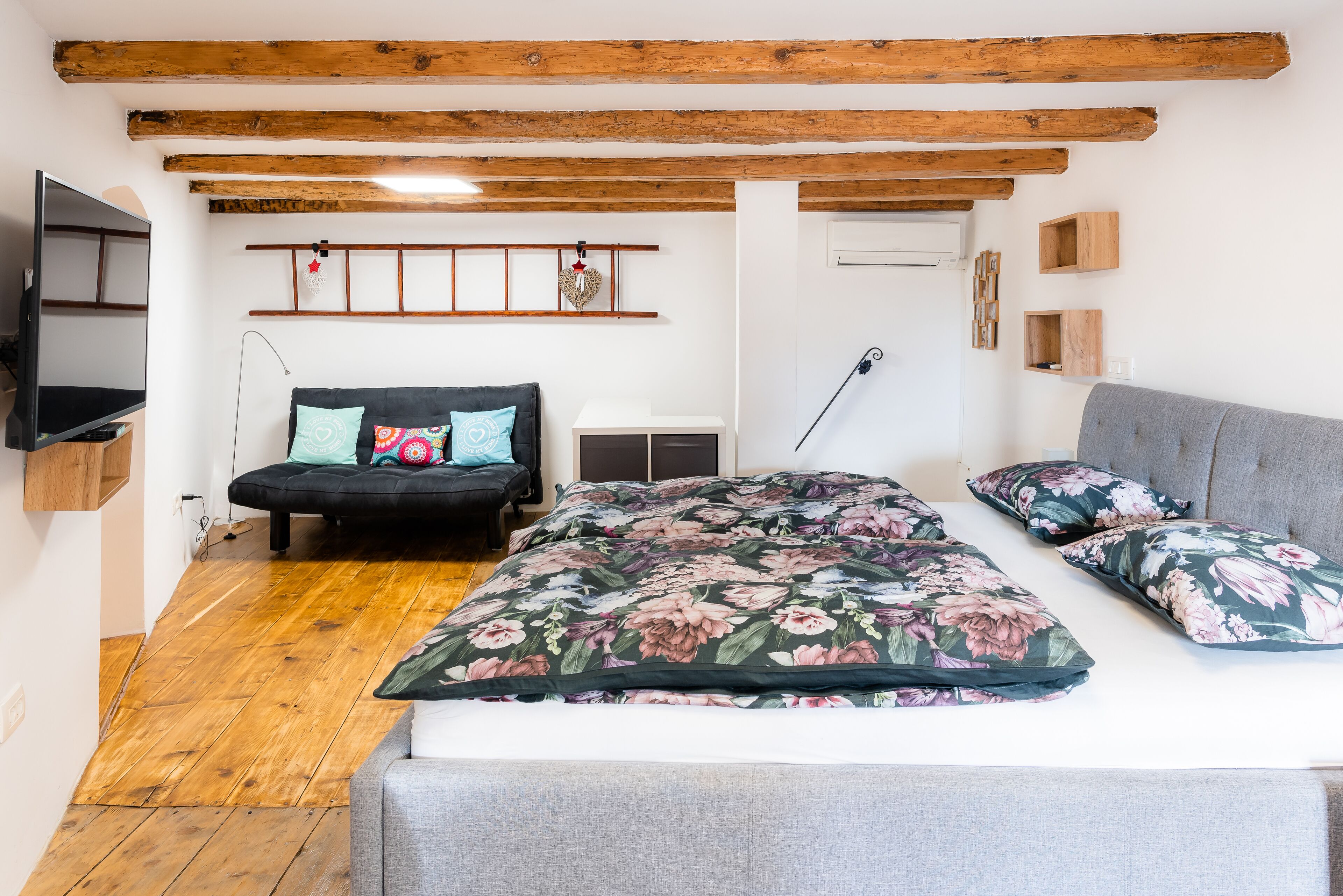 A bedroom with a bed, a sofa, wooden beams on the ceiling, a TV, and a wall-mounted shelf.