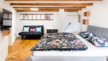A bedroom with a bed, a sofa, wooden beams on the ceiling, a TV, and a wall-mounted shelf.