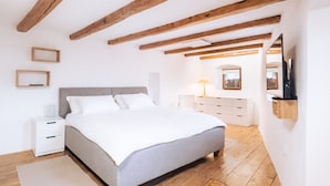 Een slaapkamer met een bed, houten balken, witte muren, een nachtkastje, een dressoir en een spiegel.