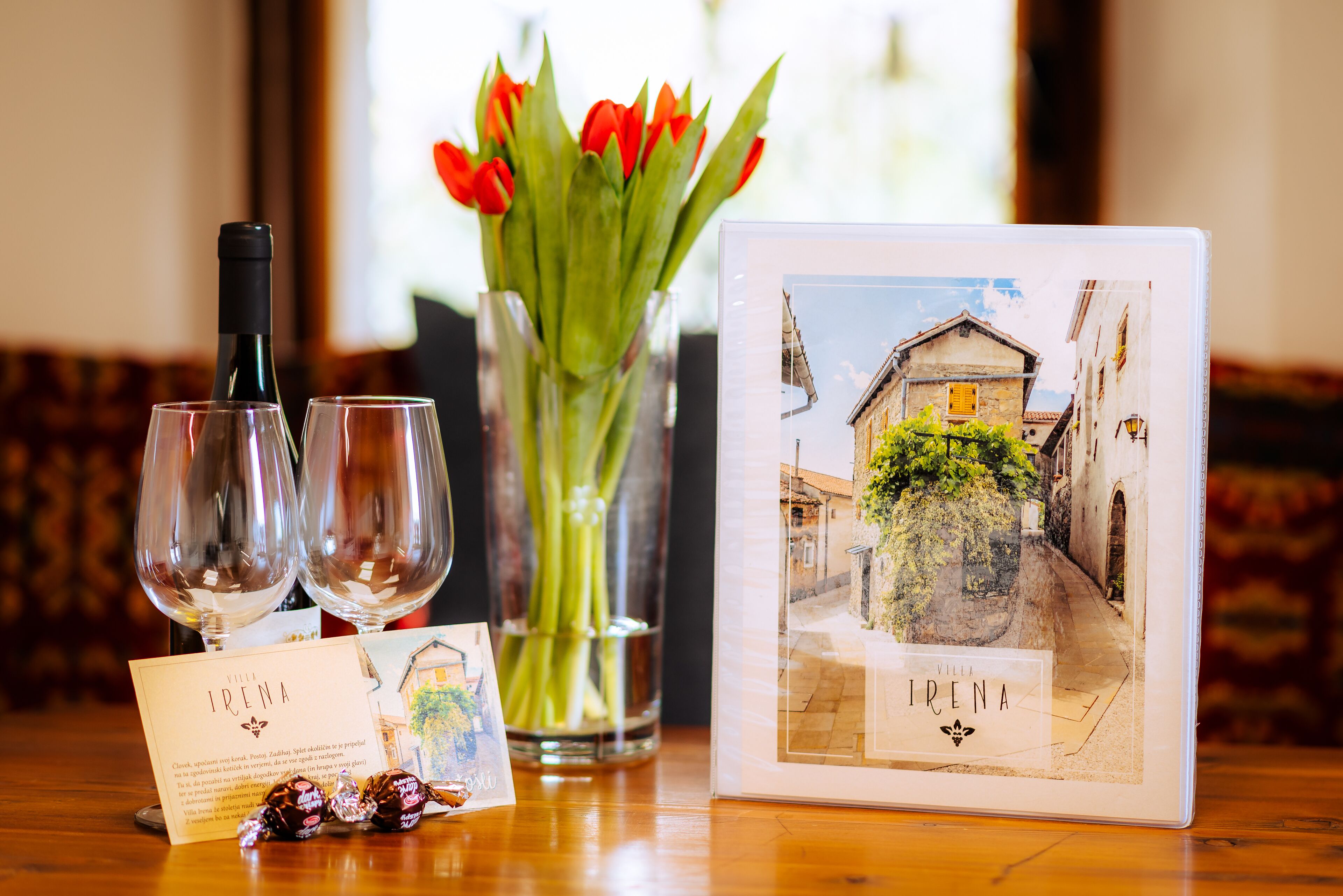 A table with a wine bottle, two glasses, a card, a framed picture, and a vase with red tulips.