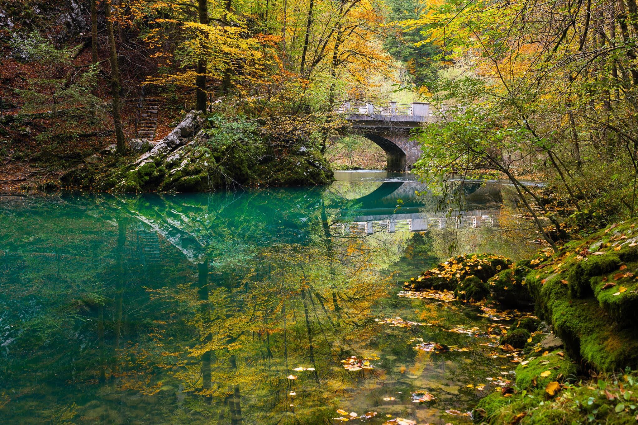 A stone bridge over a clear river surrounded by autumn foliage.