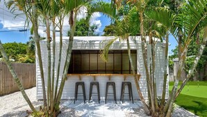 A modern outdoor bar area with a white structure, three stools, and palm trees.