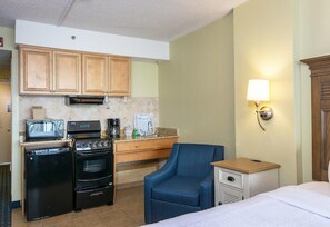 Una habitación de hotel compacta con kitchenette, una cama, una silla y una mesita de noche.