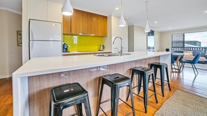 Una cocina moderna con encimera blanca, armarios de madera y salpicadero amarillo. Hay tres taburetes negros y un ventanal con vistas al exterior.