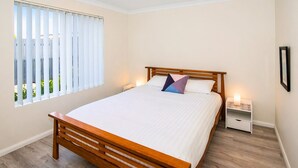 Een slaapkamer met een houten bed, wit beddengoed, een nachtkastje met een lamp en een raam met jaloezieën.