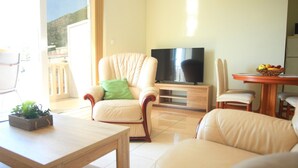 Sala de estar com sofá, poltronas, mesa de centro, televisão e mesa de jantar com frutas.