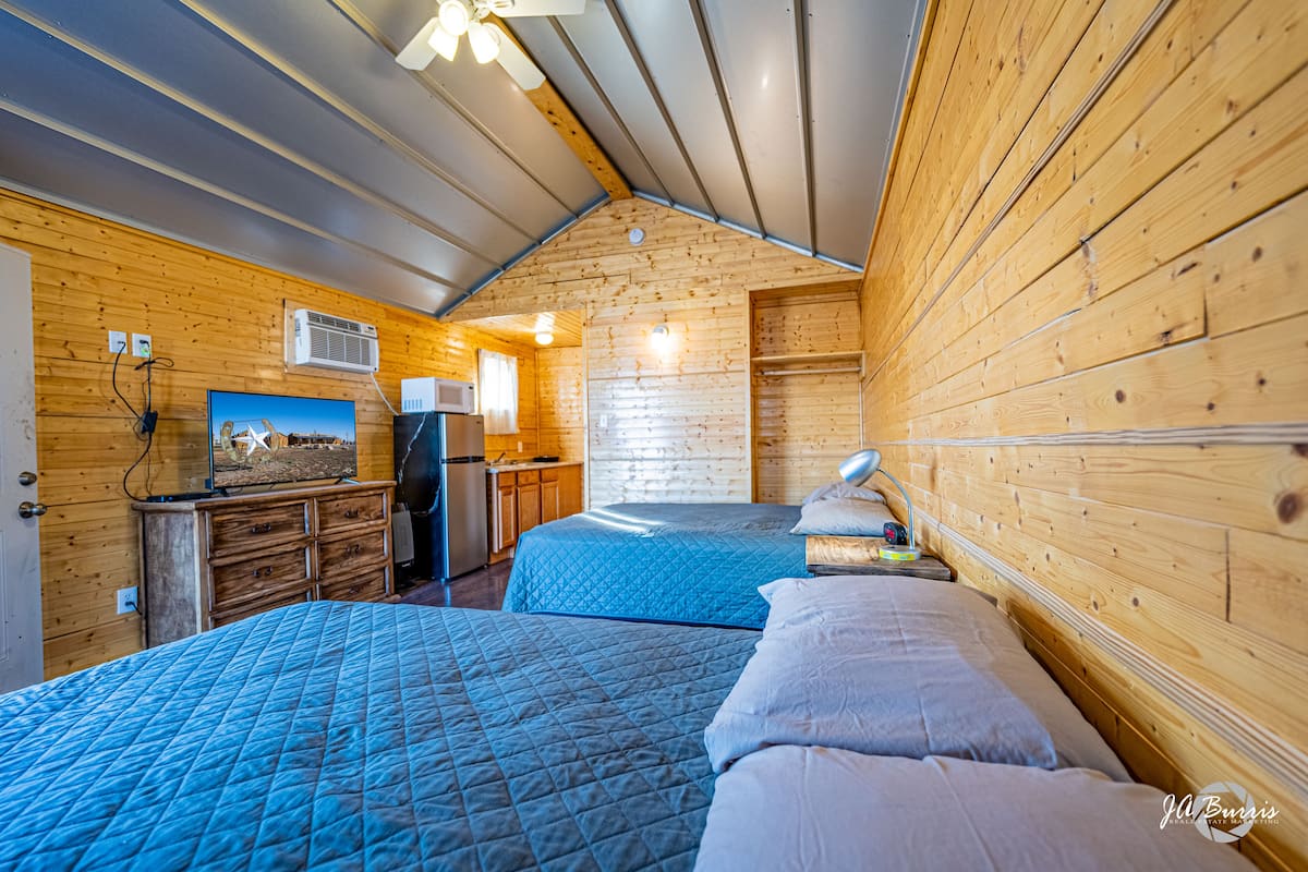 A cabin with two beds, wooden walls, a ceiling fan, a kitchenette, and a mounted TV.
