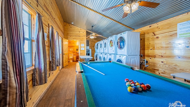 Une table de billard dans une pièce aux murs et au plafond en bois, avec un ventilateur de plafond et une rangée de machines à laver.