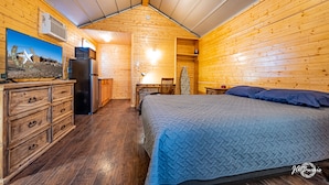 Une chambre confortable, aménagée dans le style d’un chalet, avec un intérieur en bois, un lit recouvert d’une courtepointe bleue, une commode, un réfrigérateur, un bureau avec une chaise et un téléviseur fixé au mur.