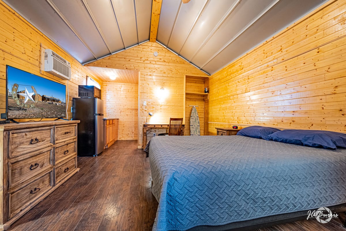 A cozy cabin-style room with a wooden interior, a bed with a blue quilt, a dresser, a refrigerator, a desk with a chair, and a mounted TV.
