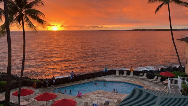 Un espace piscine offrant une vue sur le coucher de soleil, avec des palmiers et des chaises longues.