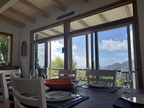A dining area with a table set for two, large windows offering a view of mountains and greenery, and a ceiling with wooden beams.
