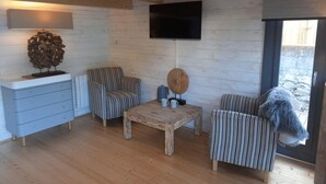 A cozy room with a wooden ceiling, a flat-screen TV mounted on the wall, a wooden coffee table, striped chairs, and a small side table with a decorative object. - Sandy Toes Accommodation (Par)