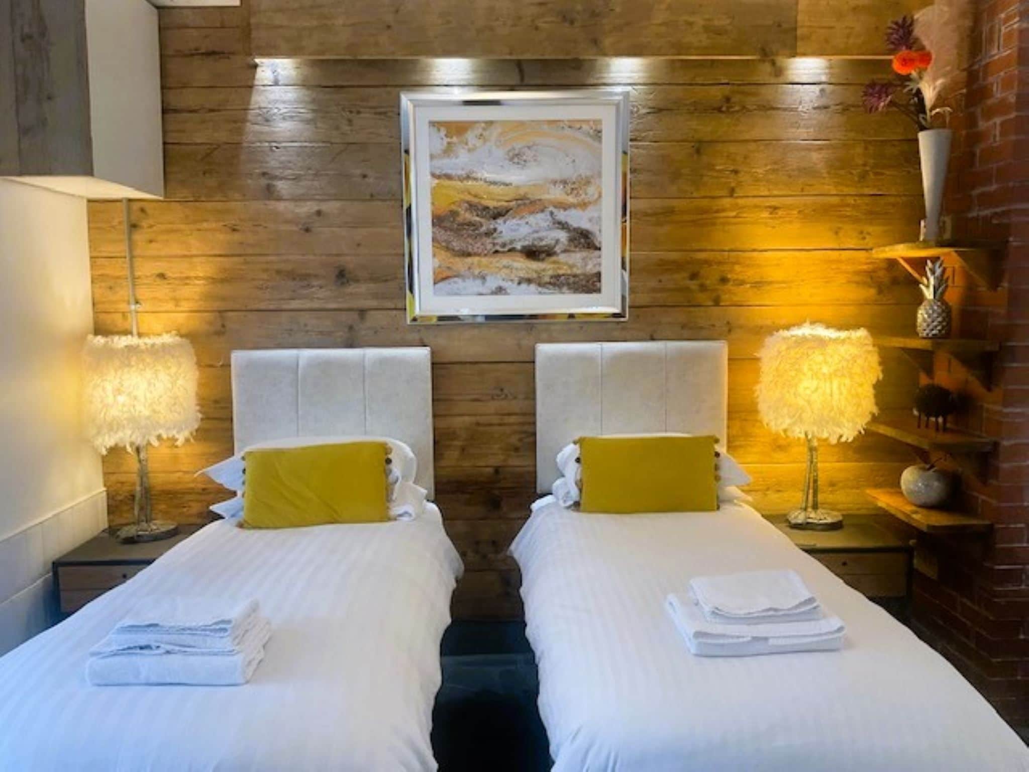 A room with two beds, each with white bedding and a single yellow pillow. There is a framed painting above the beds, and a lamp on each bedside table.