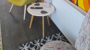 A modern living room with a yellow armchair, a small round table with a remote, and a vase with flowers. A TV is mounted on the wall, and the room has a vibrant orange and yellow patterned wallpaper. - Sandy Toes Accommodation (Par)