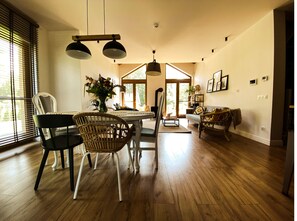 Un vaste espace de vie comprenant une table à manger, des chaises et un canapé. On y trouve une grande fenêtre avec des stores, une cheminée et un luminaire au plafond.