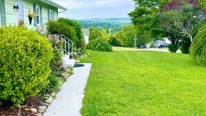 A residential property with a well-maintained lawn, a pathway leading to a house with green shutters, and a view of a hilly landscape in the background.