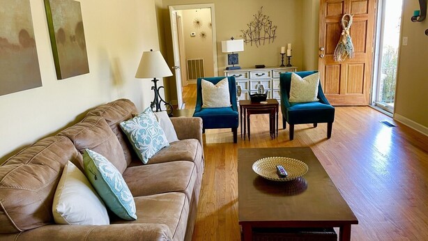 A living room with a brown leather sofa, teal armchairs, a wooden coffee table, and a dining table with a decorative bowl.