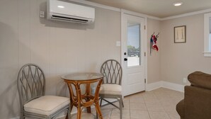 Een kamer met een glazen tafel, twee rieten stoelen, een bruine bank, een airconditioner, een deur met een raam en een lijst met een afbeelding aan de muur.