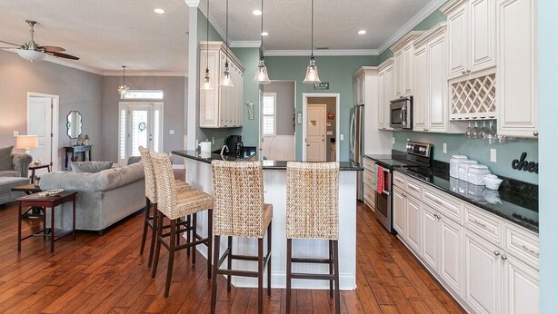 A spacious kitchen with white cabinets, a central island with bar stools, and a dining area with a gray sofa. - Treasure Beach House with private pool and dock (ST. AUGUSTINE)