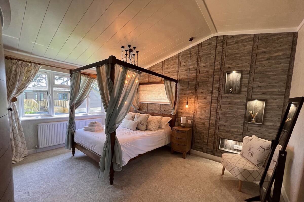 Un dormitorio con cama con dosel, mesita de noche, una silla y un ventanal con cortinas.