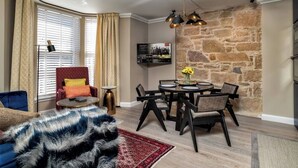 Ein Wohnzimmer mit einer Couch, einem roten Sessel, einem Couchtisch, einem an der Wand montierten Fernseher, einer Steinwand und einem Essbereich mit Tisch und Stühlen.