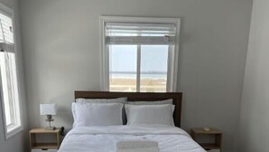 Ein Schlafzimmer mit einem großen Bett, zwei Nachttischen, einem kleinen Tisch und einem Deckenventilator. Ein Fenster bietet Blick auf den Strand.