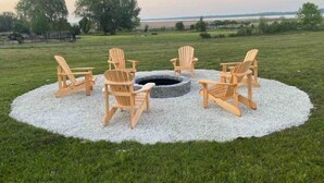 Ein kreisförmiger Sitzbereich mit Holz-Adirondack-Stühlen um eine Steinkaminfeuerstelle, auf einem Kiesrondell, inmitten eines grünen Rasenfelds mit Bäumen und weiter Aussicht.