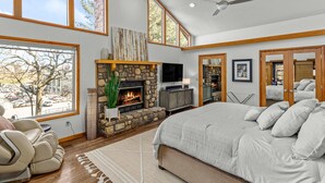 A spacious bedroom with a large bed, a stone fireplace, a television, and a view of a parking lot through large windows.
