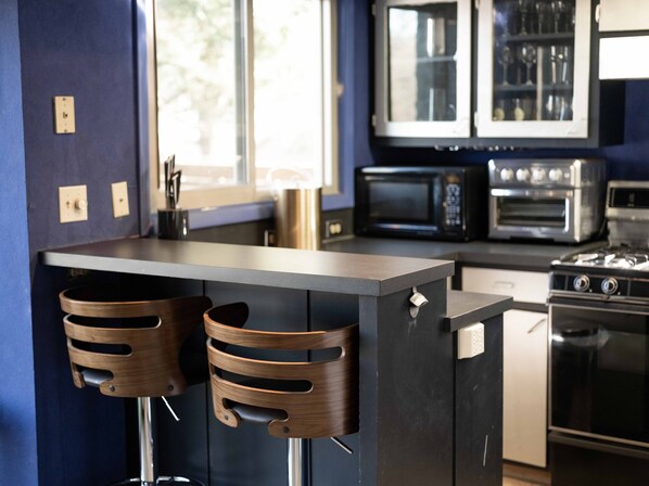 A modern kitchen with a dark countertop, two wooden bar stools, a microwave, a toaster, and a stove. - Luxury home overlooking Razorback Stadium, Old Main and Alumni House. (Fayetteville)
