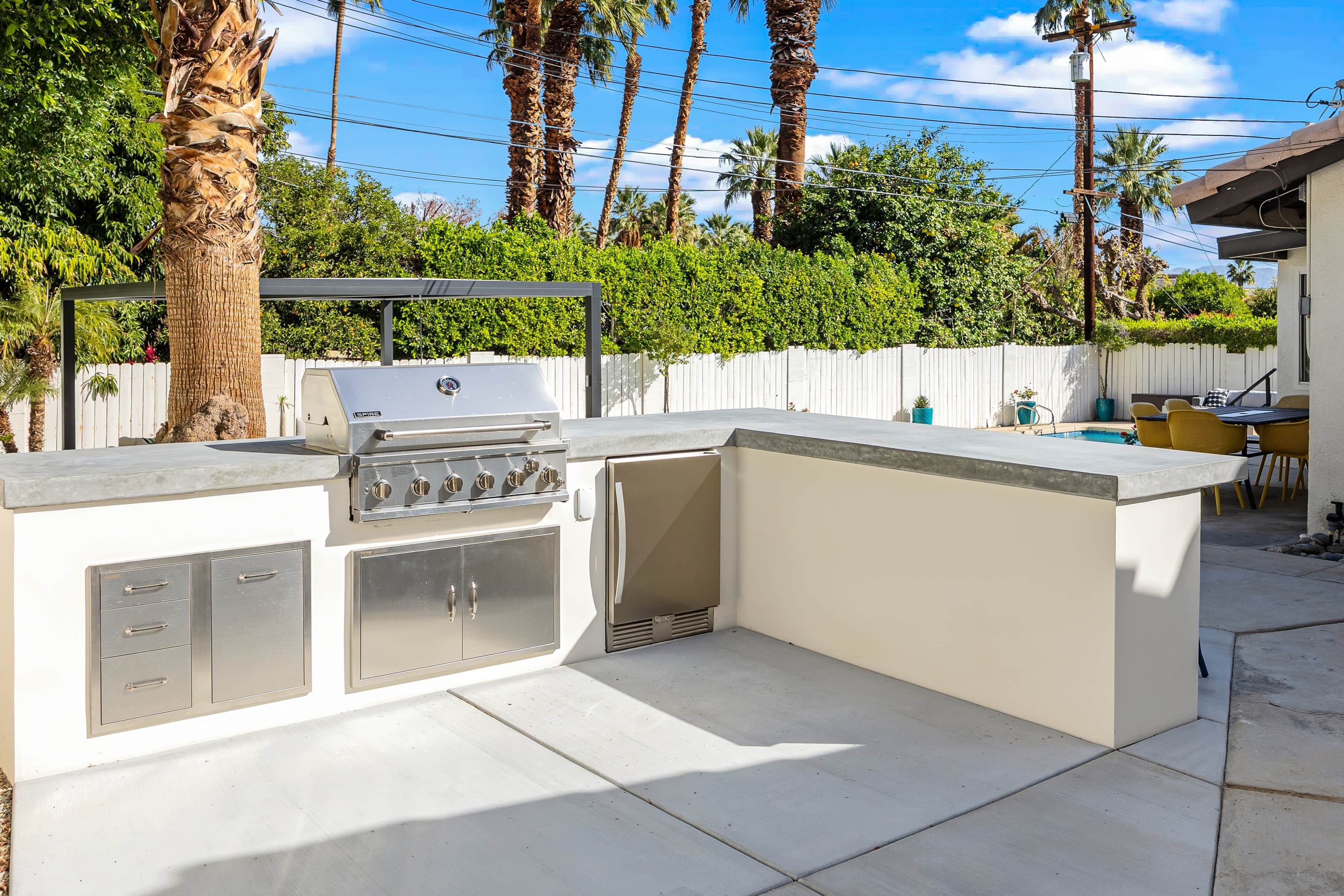 Une cuisine extérieure équipée d’un barbecue, d’un évier et de placards de rangement, entourée d’une clôture blanche et de palmiers.