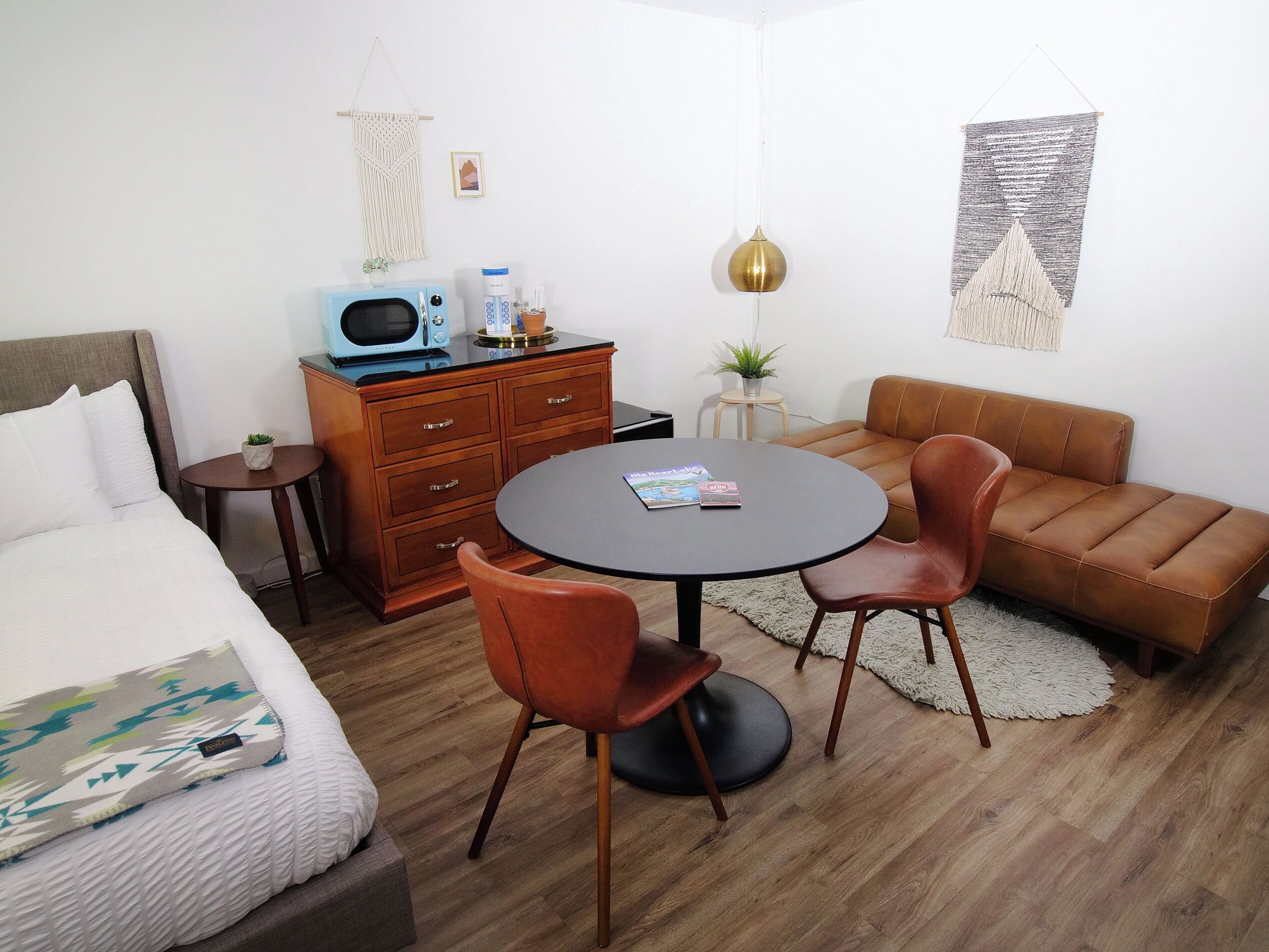 A compact hotel room with a bed, a small table with chairs, a sofa, a microwave, a small plant, and a hanging light fixture.