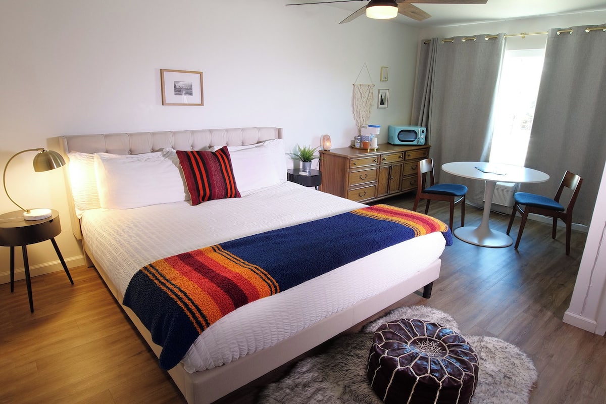 A neatly arranged bedroom with a bed, bedside lamps, a dresser, a microwave, a chair, and a small table.