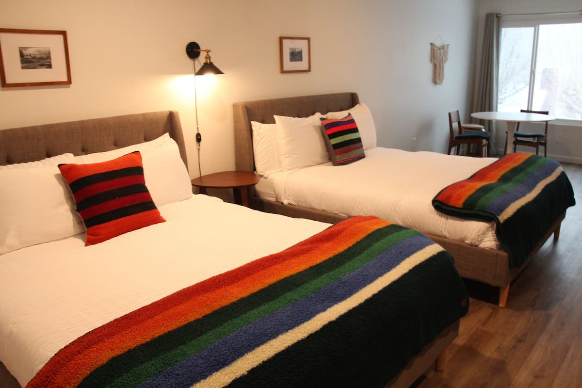 Two single beds with white linens and colorful striped pillows, a small round table, and a chair in a room with a window and framed pictures on the wall.