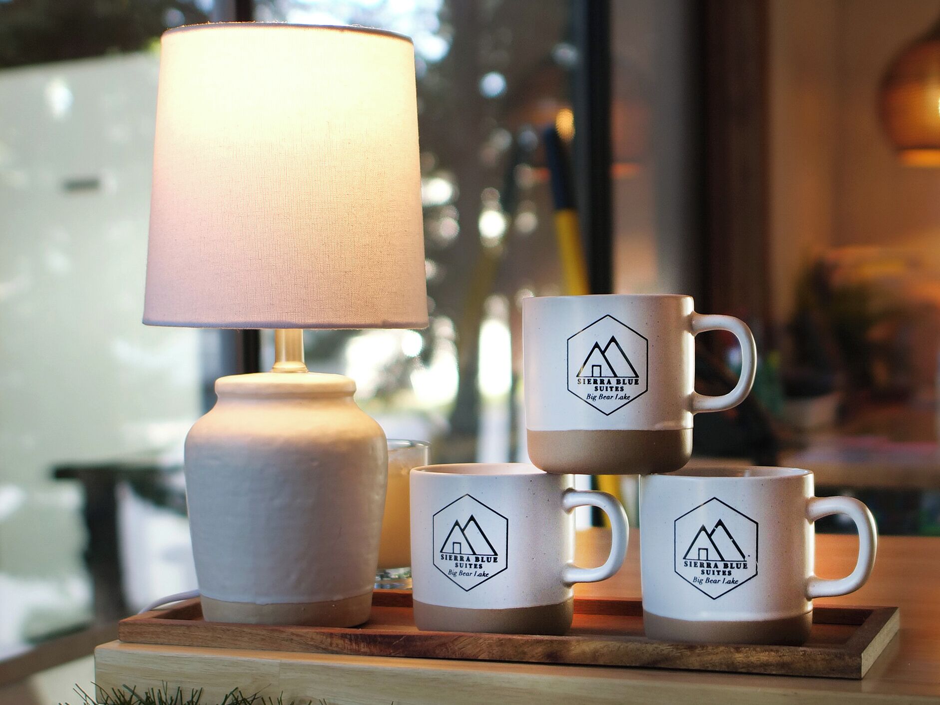 Une table avec une lampe et trois tasses ornées d’un logo représentant une montagne, une bougie et un verre d’eau.
