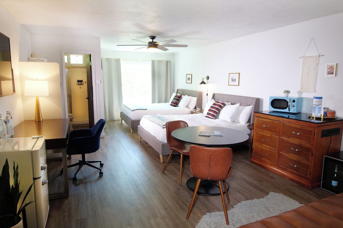 A hotel room with two beds, a dining table, a desk, a chair, a ceiling fan, and a microwave.