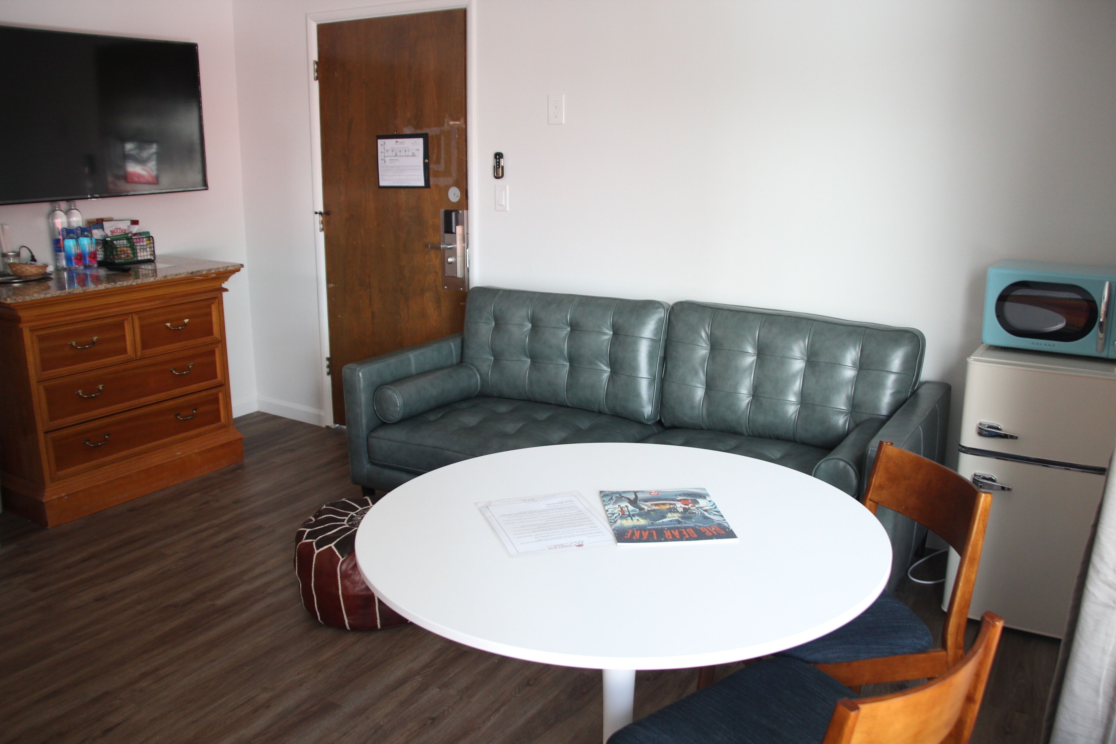 A compact living space with a round white table, a green leather sofa, a wooden dresser, a television, and a small refrigerator.