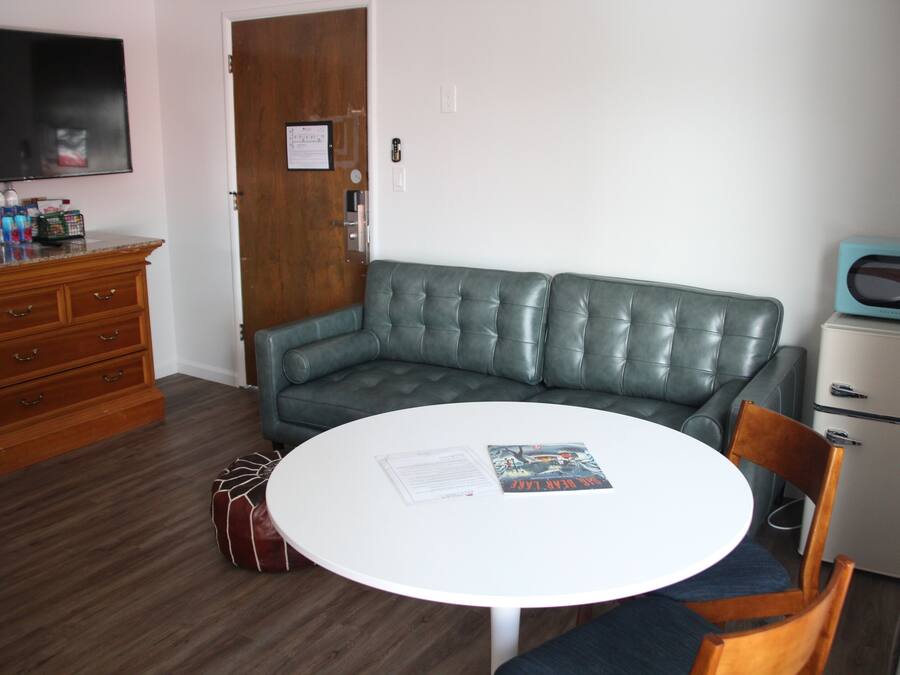 A compact living space with a round white table, a green leather sofa, a wooden dresser, a television, and a small refrigerator.