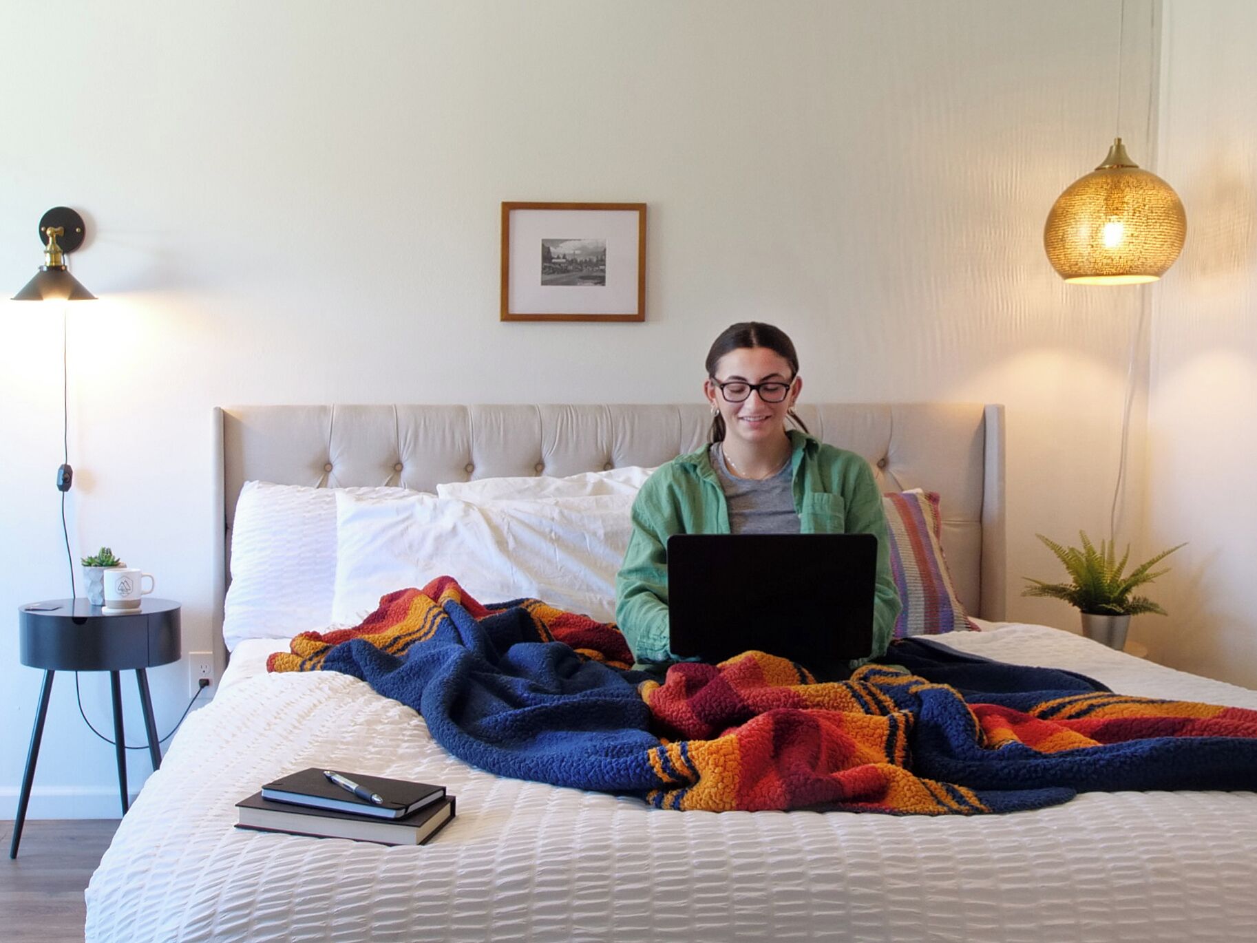 A person sitting on a bed with a laptop, a colorful blanket, and a framed picture on the wall.
