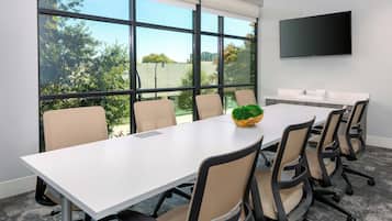 A conference room with a large table, six chairs, a TV on the wall, and a view of trees and buildings outside.