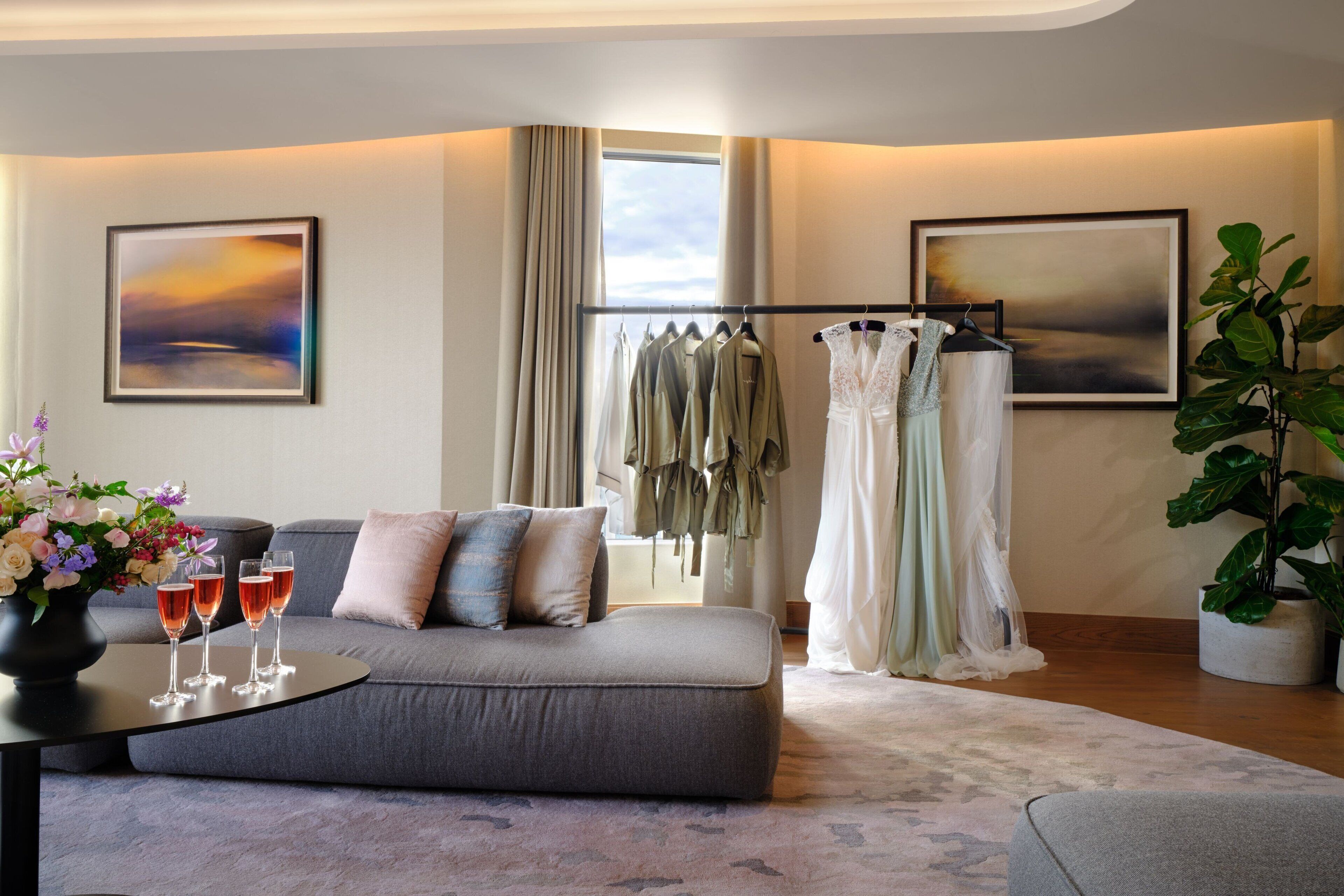 Ein modernes Hotelzimmer mit einer grauen Couch, einem Couchtisch mit Getränken, einem Kleiderständer und zwei großen Landschaftsbildern.