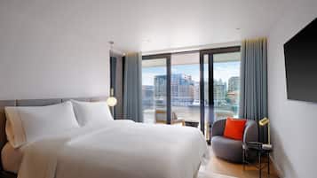 A modern hotel room with a large bed, a seating area with a chair and a small table, and a large window offering a city view.